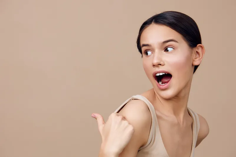 Excited young woman with a cheerful expression against a neutral beige background, showcasing joy and enthusiasm in a stylish casual outfit