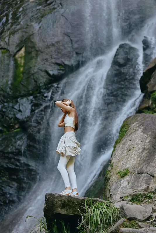 woman standing near waterfall wearing white activewear, surrounded by rocks and greenery, enjoying nature outdoors in a peaceful moment.