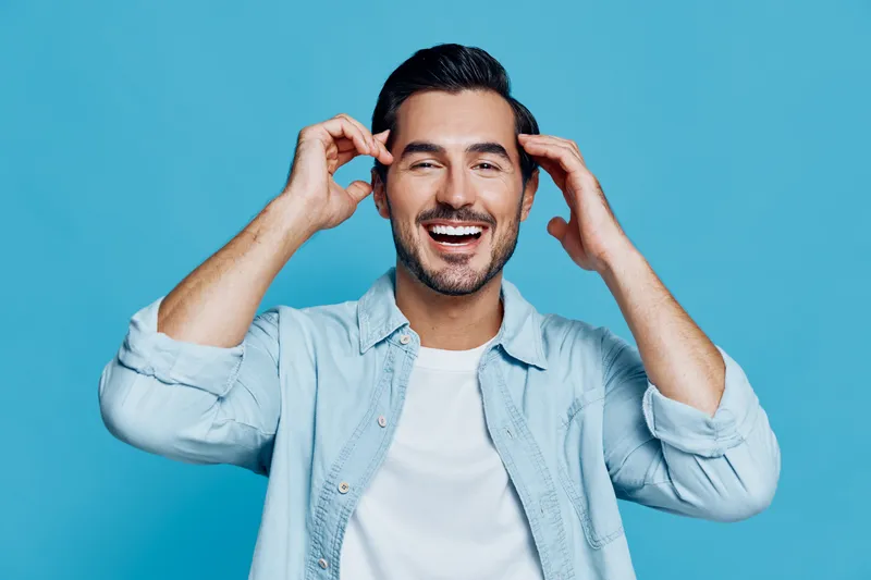 Smiling happy cheerful young man wearing casual clothes poses with hands on his head against a plain solid blue background, expressing joy and positivity.
