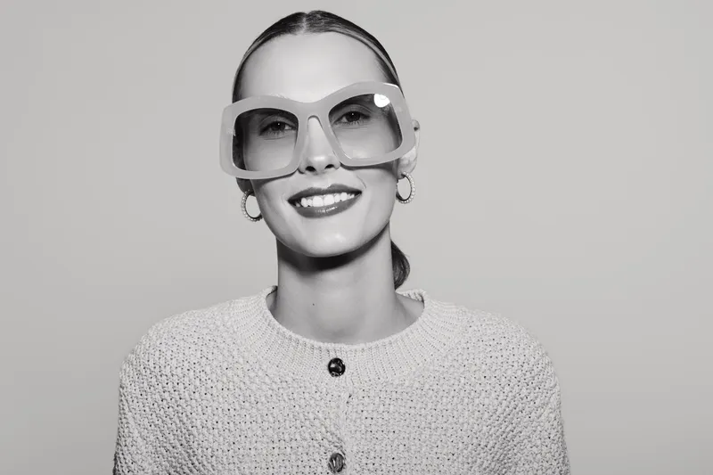 Smiling caucasian woman with pale skin and slicked hair wears large sunglasses and textured sweater in studio light.