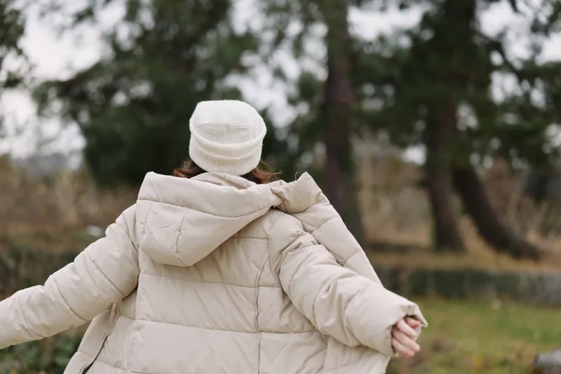 Person wearing a beige winter coat with arms outstretched in a park, outdoor scene conveys calm, freedom and cozy charm in cool weather