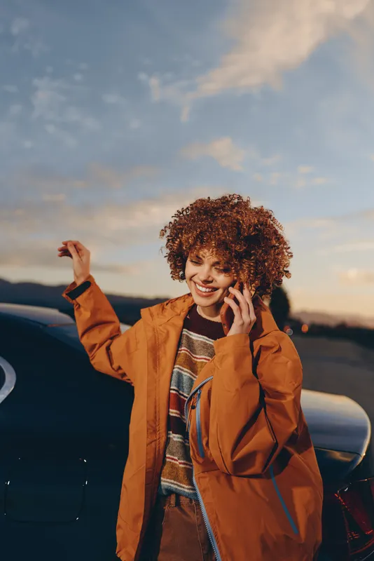 Woman lifestyle outdoors at sunset smiling and talking on phone with curly hair, wearing an orange coat near a parked car, warm sky background, relaxed evening road trip carefree moment