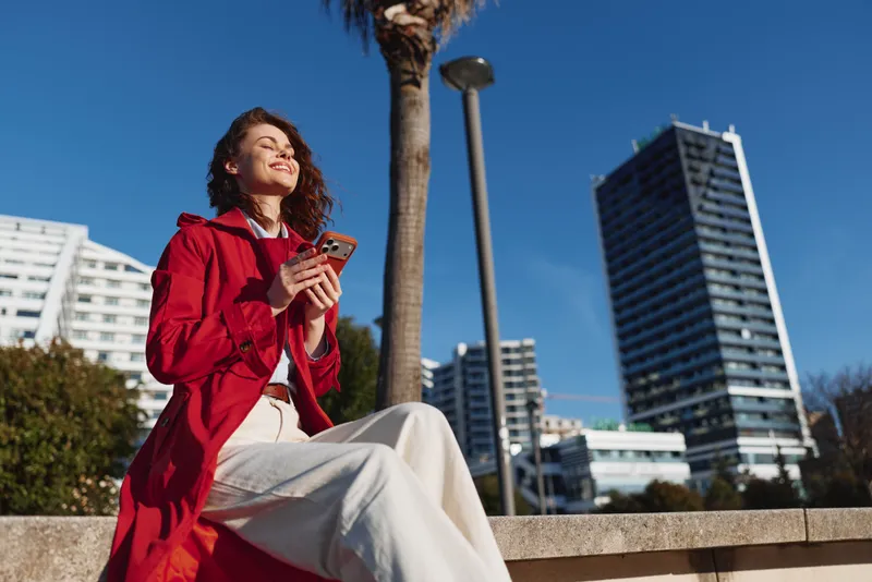 Young woman in red coat sits outdoors on concrete bench holds smartphone enjoys sunny day with modern buildings and blue sky in background urban lifestyle concept