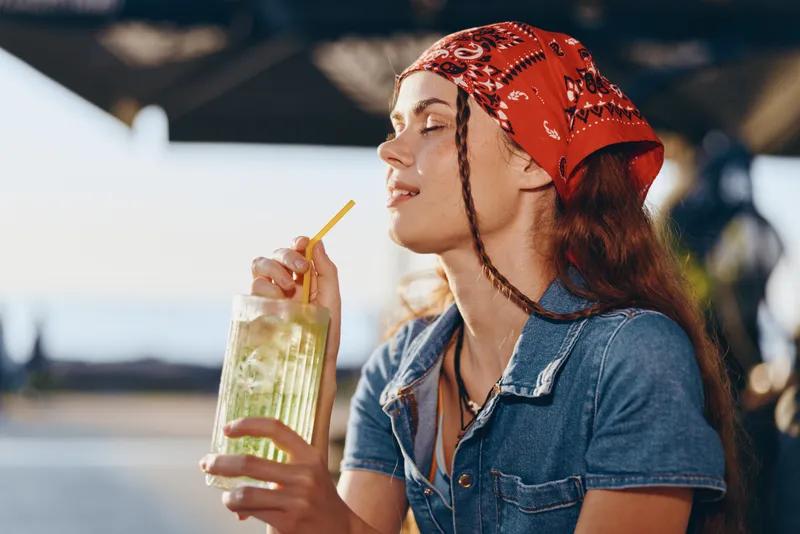 young woman enjoying refreshing summer drink wearing red bandana and denim shirt with relaxed outdoor background in warm sunlight