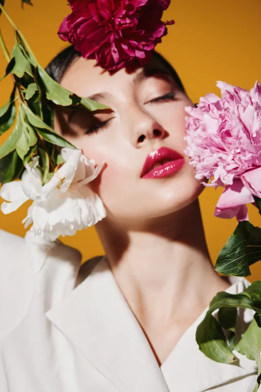 Stylish woman poses with closed eyes among vibrant pink white and red flowers on mustard background wearing white blazer. Concept of beauty fashion creative and floral elegance.