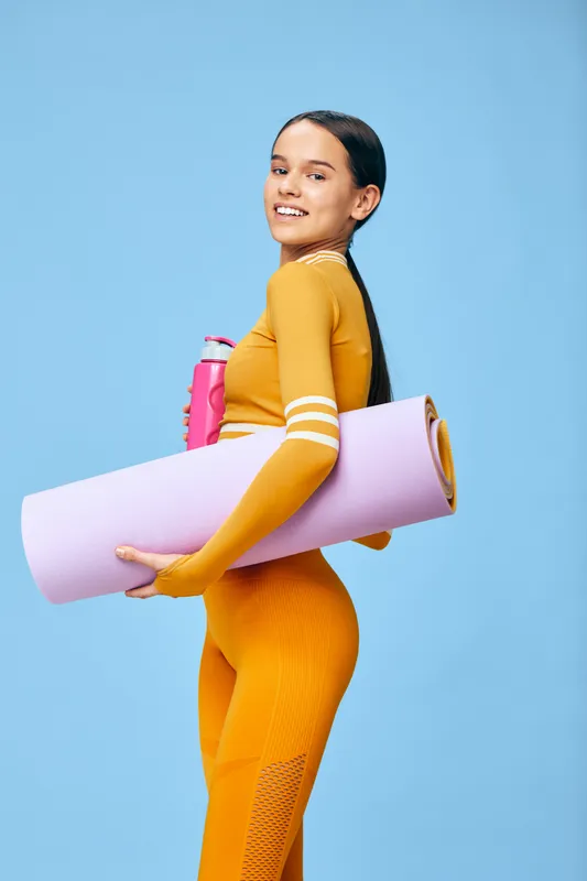Woman in vibrant athletic wear carries a yoga mat and water bottle, ready for a workout and sport session. Bright studio backdrop highlights health, training and athletic lifestyle as she prepares