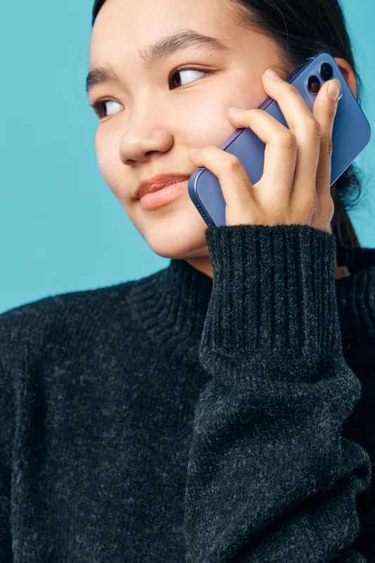 Young woman in black sweater holding smartphone to ear and looking aside on blue background for communication conversation or call with mobile device female user technology and connection
