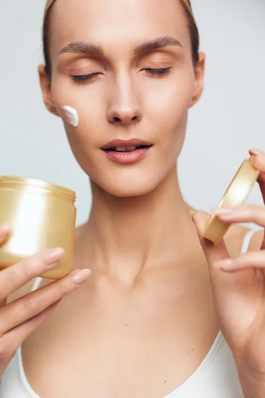 A young woman applies cream from a gold container on her face, standing against a neutral background, her calm expression conveying a gentle skincare moment.