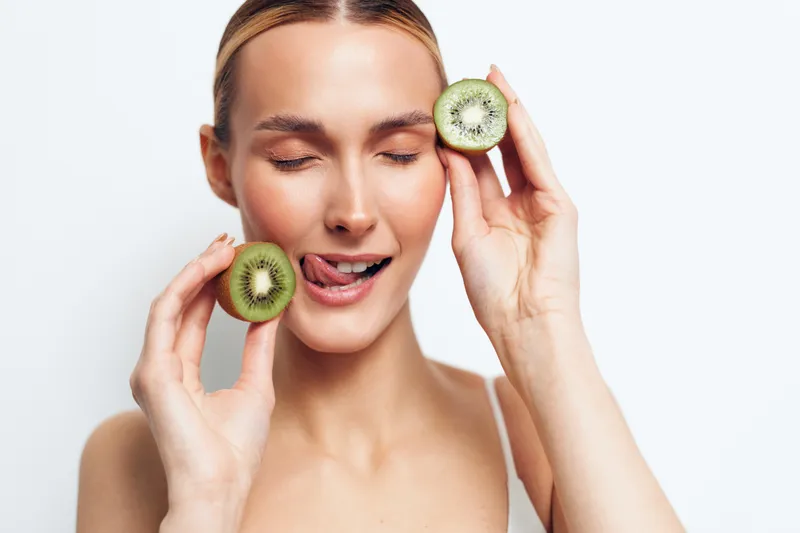 A young woman with closed eyes holds two halves of fresh kiwi fruit near her face while smiling slightly and licking her lips, representing healthy lifestyle and natural beauty concept.