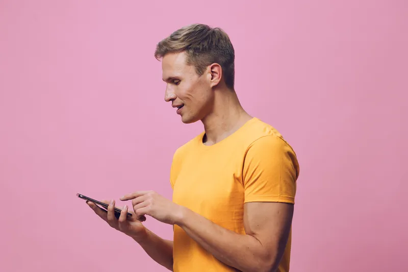 smiling young man using smartphone wearing yellow t-shirt isolated pink background digital nomad chat with AI concept