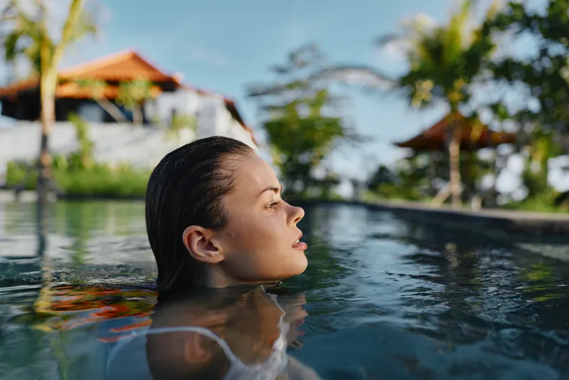 Close-up of a woman with wet hair in a pool, side profile, sunny tropical backdrop. Relaxation, leisure, vacation vibes, calm water, outdoor scene, warm breeze, water reflections, leisure lifestyle.