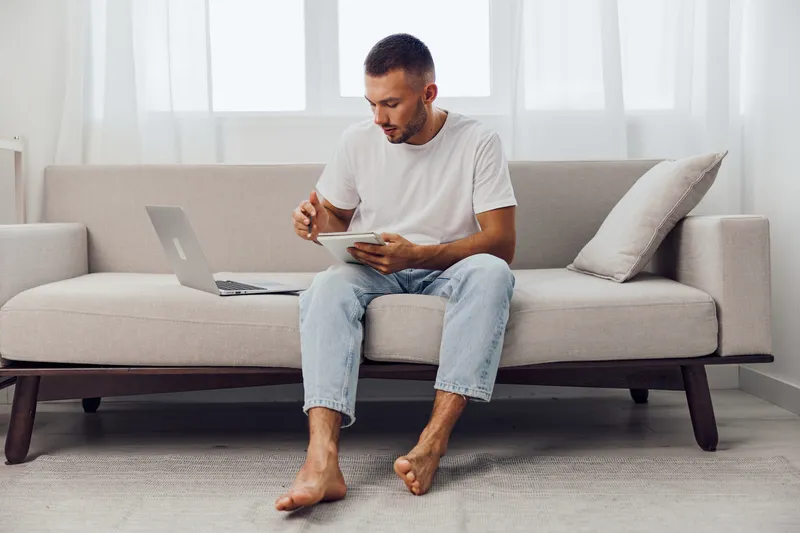 Focused man working on a tablet while sitting on a modern couch, wearing casual attire in a bright, airy room that reflects contemporary home office trends.