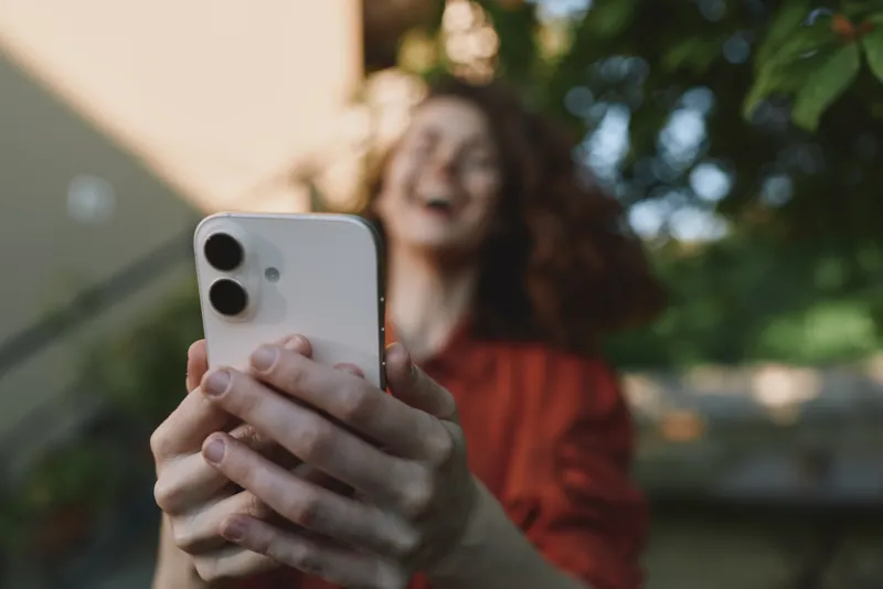 Cheerful woman outdoors, smiling while taking a selfie with a smartphone, casual lifestyle moment, sunlit garden background, relaxed pose, natural colors