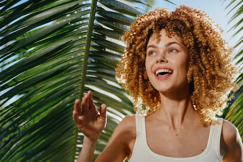 Smiling woman with curly hair enjoying sunny tropical outdoors surrounded by green palm leaves, wearing a white tank top, expressing happiness and natural beauty.