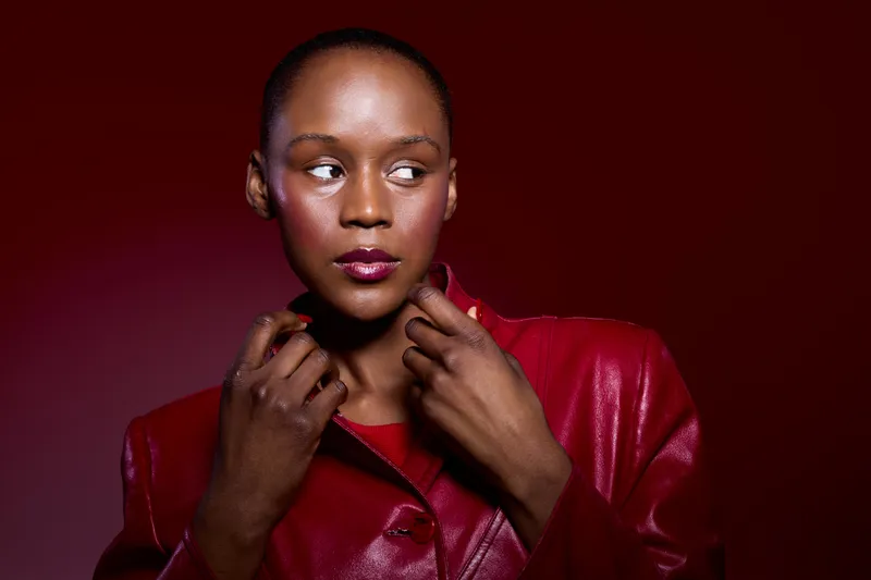 Portrait of a dark skinned African woman wearing a shiny red jacket, looking to the side on a red background. Concept of fashion, beauty, confidence, and elegance in studio lighting.