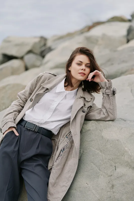Young woman in stylish coat and white shirt leans on rock outdoors. Casual fashion portrait with natural background and relaxed confident pose. Concept of modern urban style and elegance.