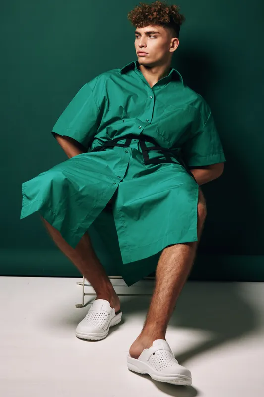 Young man sitting on chair wearing green oversized shirt dress with black belt and white slip on shoes against dark green background in studio.