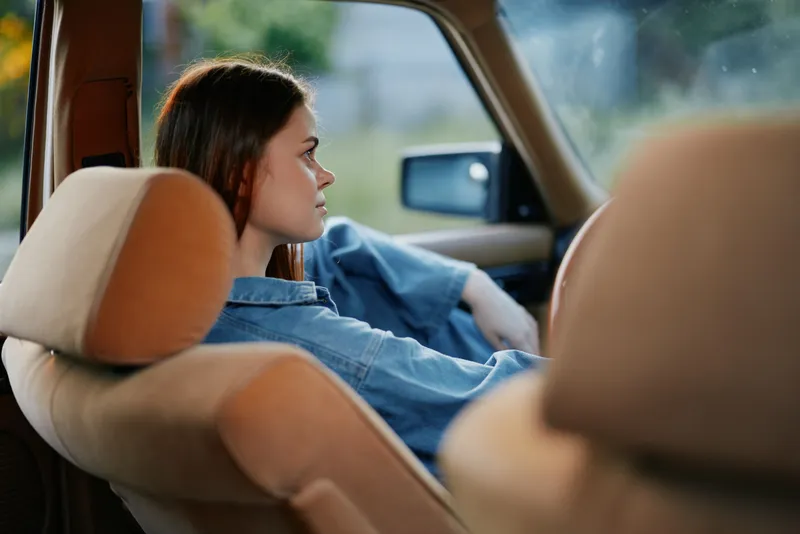 Young woman in casual denim outfit reflecting in car interior, showcasing a sense of calm and contemplation in natural light.