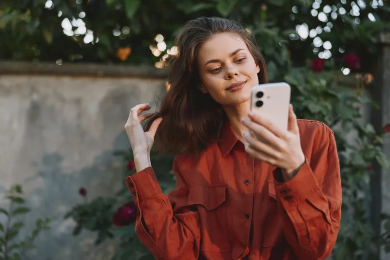 Bright outdoor portrait of a woman in a casual red shirt, taking a selfie, relaxed pose, sunlit greenery background, natural tones, candid lifestyle moment