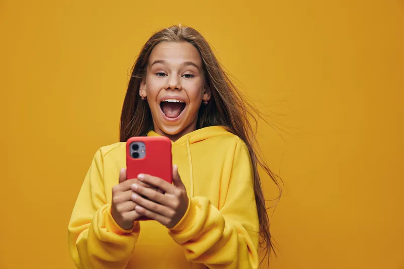 Happy girl in yellow hoodie holding red smartphone and looking excited with open mouth on bright yellow background. Casual lifestyle and positive emotions in portrait.