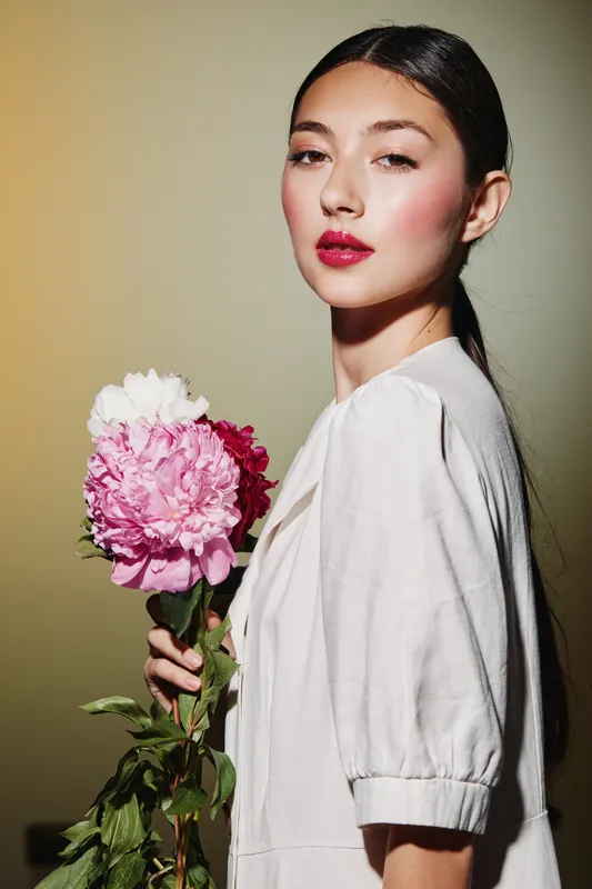 Young woman with pink makeup holding multi-colored peonies posing creatively against light gradient background in white blouse. Concept for beauty, fashion, and floral elegance.