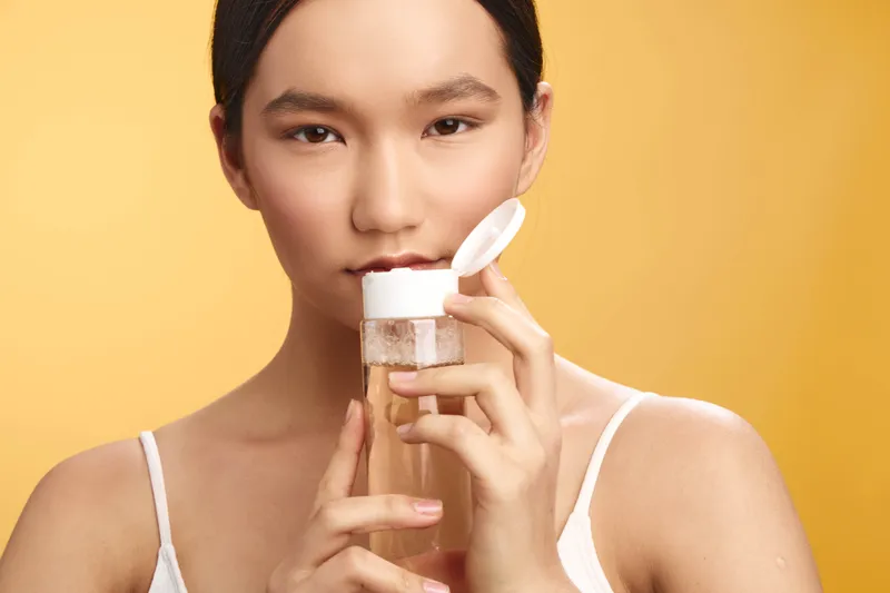 asian woman applies skincare product in a bright studio, showcasing a fresh complexion and a calm smile as she looks at the camera, conveying self care and beauty routines.