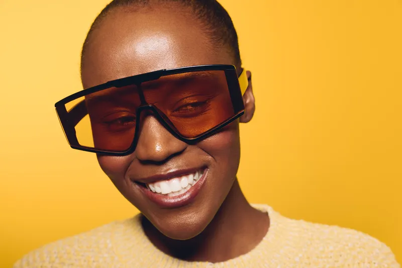 Happy African woman with dark skin wearing oversized sunglasses smiles with closed eyes against yellow background. Concept of joy, fashion, beauty, and summer lifestyle portrait.