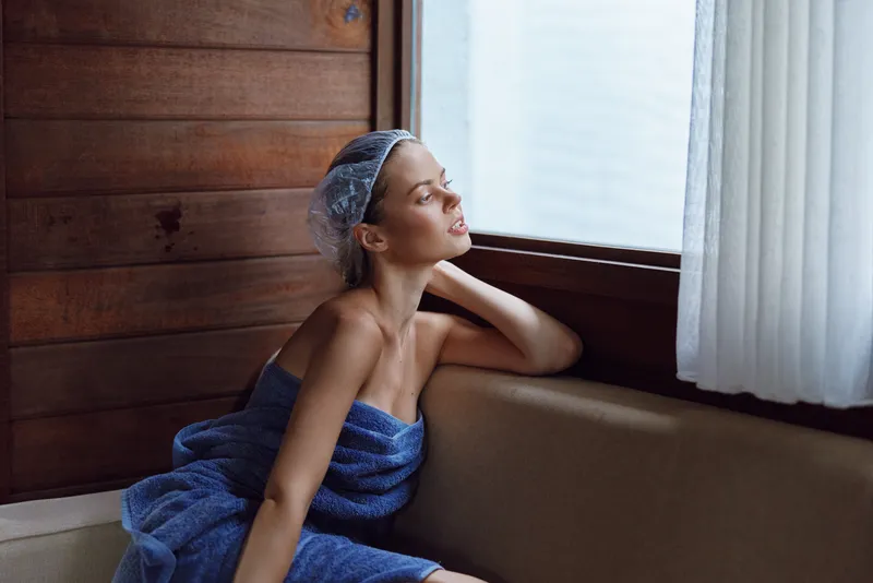 young woman wrapped in blue towel relaxing near window in wooden interior, peaceful moment with natural light illuminating soft textures and calm mood