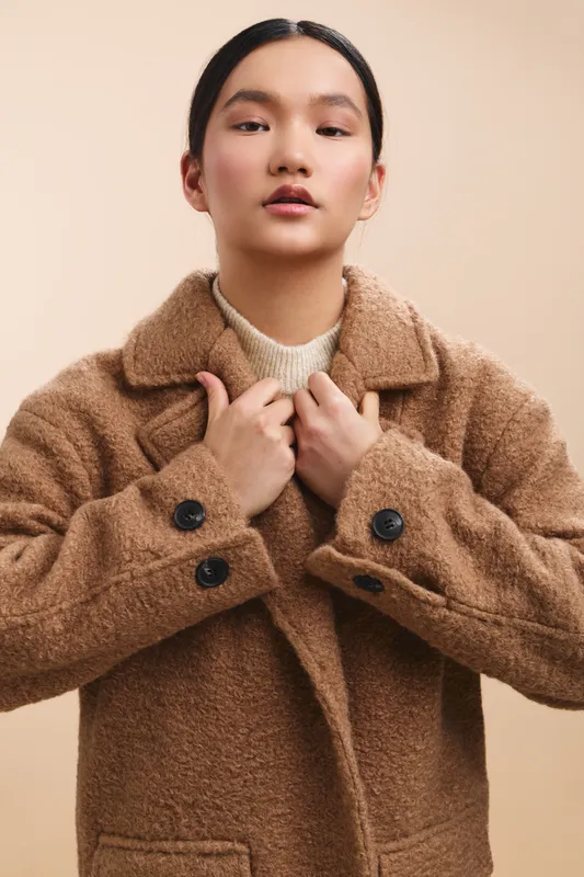 asian woman wearing a plush brown coat stands in a neutral studio, adjusting her lapels with calm confidence. her poised expression radiates warmth and modern fashion sense.
