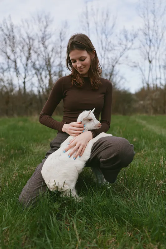 Warm outdoor moment with a calm woman kneeling in grass, softly stroking a white goat kid, gentle connection, natural light, earthy tones, serene countryside mood