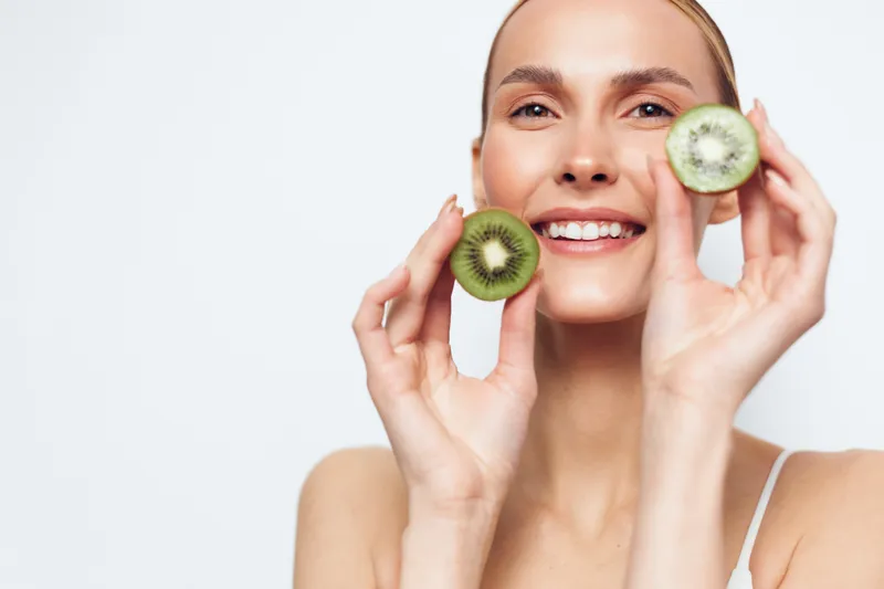 Young woman smiles brightly while holding kiwi slices near her face, posing in a clean studio for a fresh healthy lifestyle concept with a light background.
