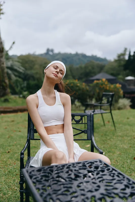 Young woman in white sportswear relaxing outdoors, enjoying nature and tranquility, featuring a peaceful expression, green grass, and a soft sky backdrop