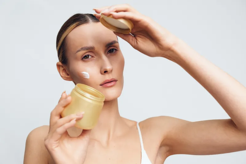 Young woman applies face cream on clean skin holds jar of moisturizer skincare beauty routine isolated on white background closeup portrait natural look.