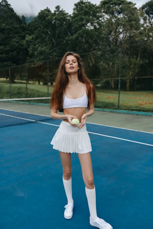Young female tennis player in sporty outfit posing confidently on blue court, exuding energy and athleticism with natural background and vibrant greenery