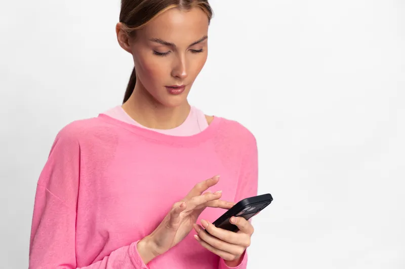 Young woman in pink sweater uses smartphone with focus and attention. Isolated on white background. Concept for communication, technology, and modern lifestyle with casual clothing.