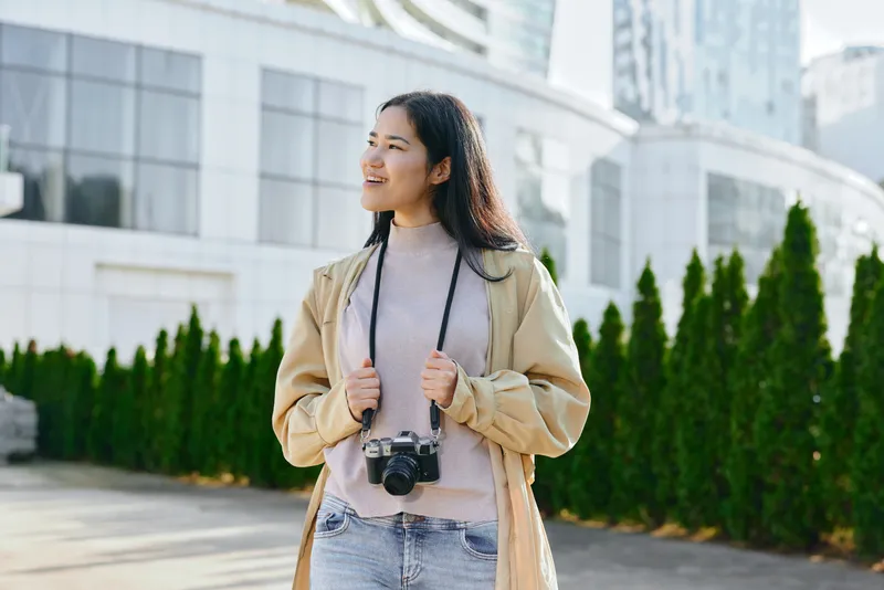 Woman with camera explores outdoor urban setting, dressed in casual fashion, smiling as she enjoys a sunny day amid modern architecture, street scenery, and relaxed travel vibe