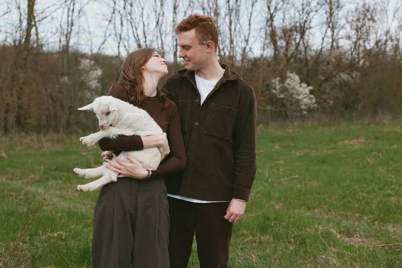 Smiling couple in casual outfits outdoors hold a playful goat kid in a grassy field, showcasing affection, nature, and lifestyle moments under warm, soft daylight and warm connection.