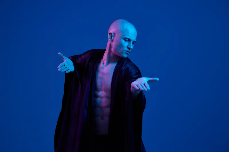 A striking man poses gracefully in a dramatic black outfit against a vibrant blue background, exuding confidence and artistry in this captivating photo.