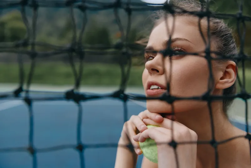 Young female tennis player with a contemplative expression, holding a tennis ball, behind a net, showcasing determination and focus in a sports setting