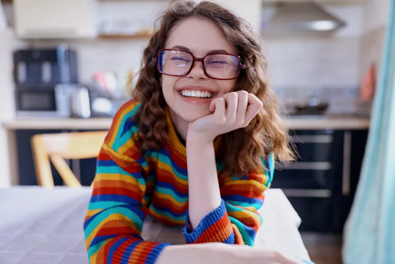 Smiling woman with curly hair wearing a bright striped sweater sits at a kitchen table, resting her chin on her hand. Cozy home lifestyle scene with warm lighting and relaxed mood Warmth.