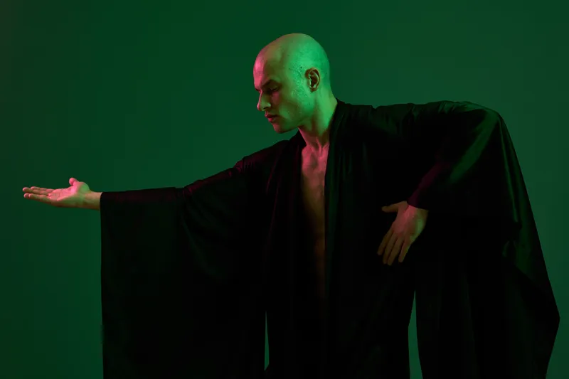 A man in a flowing black robe poses dramatically against a green background, showcasing an expressive gesture that evokes emotion and intrigue.