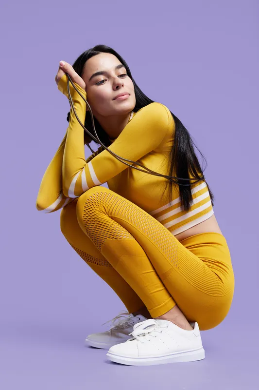 Sport fitness woman squatting in mustard yellow activewear with white sneakers, holding earphones on purple background, showing focus and determination during workout.