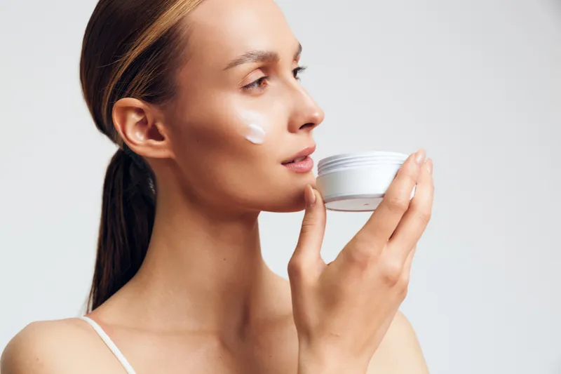 Skincare woman with face cream applying on cheek holding small jar, side profile on white background, concept of beauty wellness and selfcare, soft studio lighting, calm mood