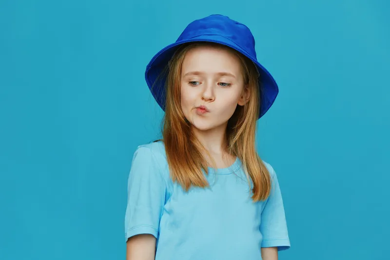 Cute girl in a blue bucket hat with long hair, showing a playful expression against a turquoise background, perfect for children s fashion and summer themes