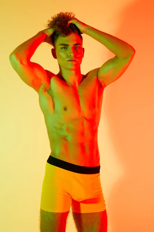 Young athletic man posing with hands on head wearing yellow shorts against colorful orange and yellow background in studio. Muscular body and confident model expression.