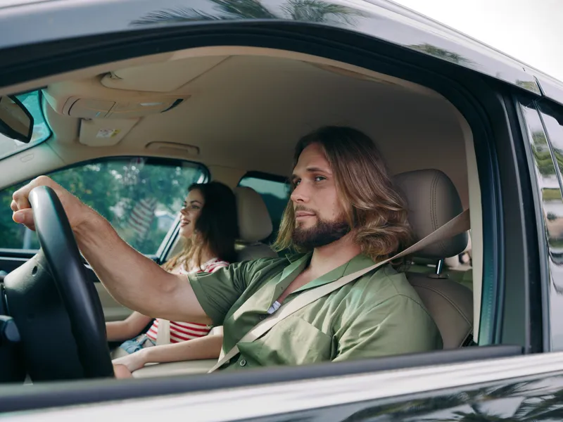 A calm man drives a car with steady hands and focused gaze while a cheerful passenger sits beside him, creating a candid daytime interior portrait of travel.