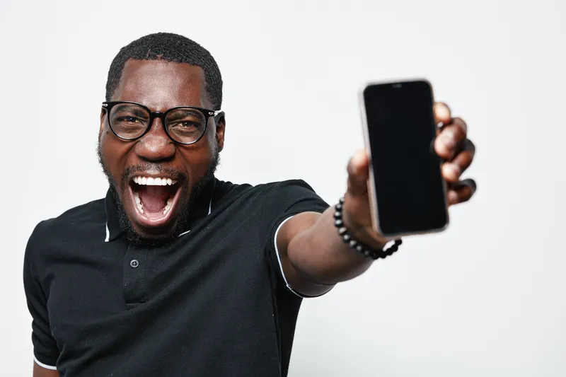 African American man with dark skin and glasses smiling excitedly while holding smartphone towards camera on white background. Happy young male showing mobile device.