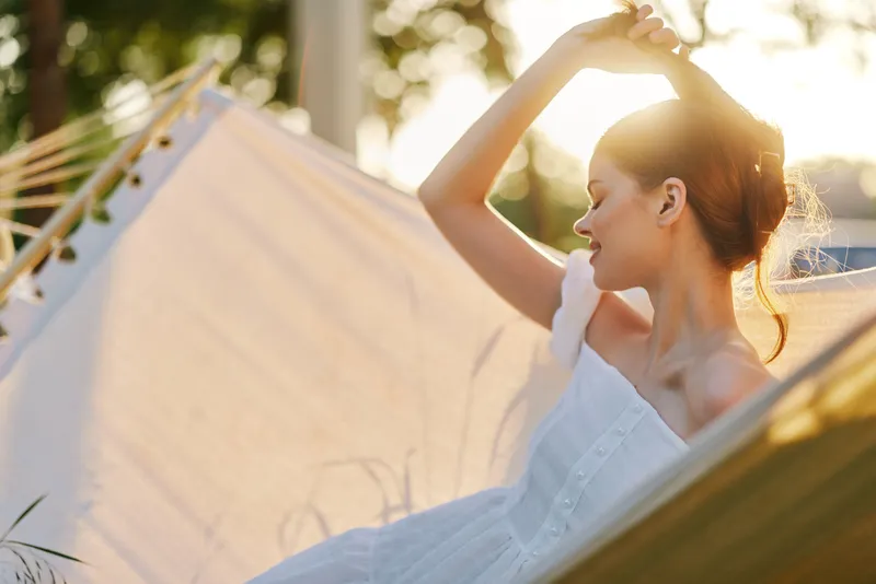 Beautiful young woman relaxing in a hammock during sunset, wearing a light summer dress, radiating calm and happiness, surrounded by a nature inspired background