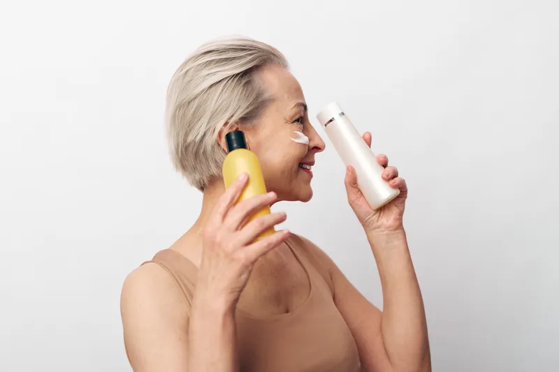 Smiling mature woman with short grey hair holds two different skincare bottles near her face on white background. Concept of beauty, skincare routine, anti aging and self care.