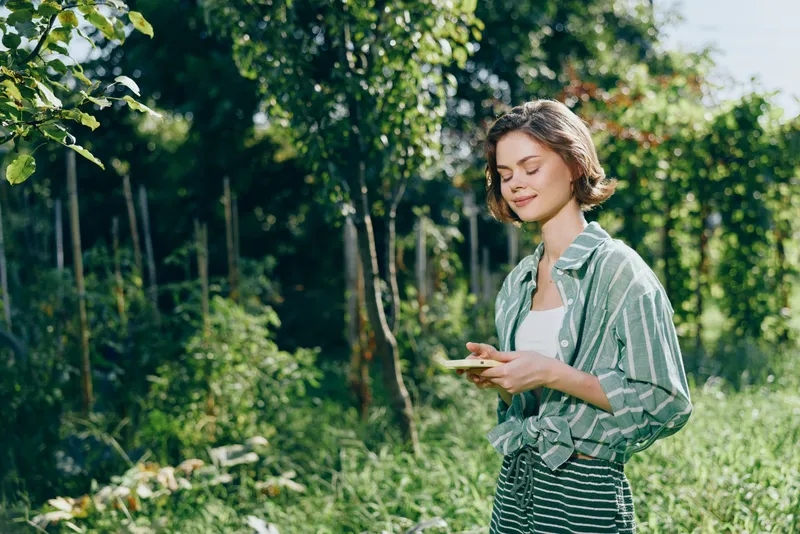 woman, outdoors, nature, calm, a cheerful woman in casual striped shirt stands among lush trees, smiling softly while looking at her phone, enjoying a peaceful moment in bright natural light and